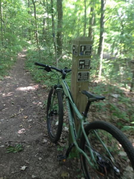 A mountain bike parked next to a trailhead sign in a lush green forest. The sign includes various trail markers and guides. A narrow dirt path leads into the trees, surrounded by dense foliage. Monte Sano State Park &amp; Land Trust mountain bike trail.