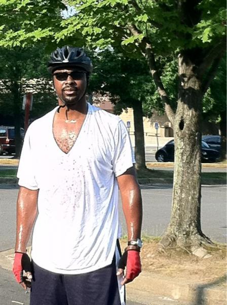 A man wearing a bicycle helmet and sunglasses stands outdoors, appearing sweaty and tired after exercising. He is dressed in a loose, white T-shirt and red gloves. In the background, there are trees and a parking area. Wakefield mountain bike trail.