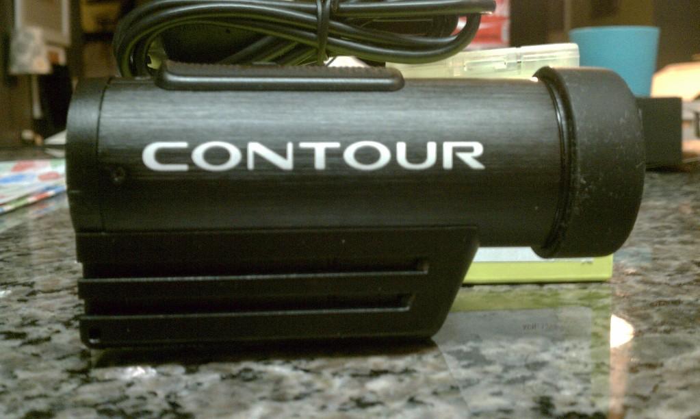 Contour Roam: Black cylindrical camera with the word "CONTOUR" printed in white on the side, resting on a granite surface. A cable is attached to the camera, and various objects are blurred in the background.