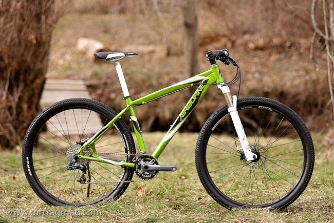 Airborne Goblin: A green mountain bike with a white seat and handlebar accents, positioned on grass near a wooded area. The bike features thick tires and a sturdy frame, suitable for off-road riding.