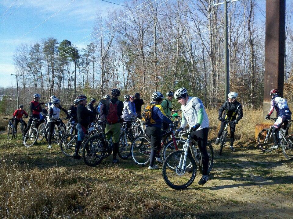 A group of mountain bikers gathered in a wooded area, standing next to their bikes on a dirt path. The scene captures a mix of cyclists wearing helmets and various outdoor gear, with trees and power lines in the background under a clear blue sky. The atmosphere suggests a friendly gathering or briefing before a ride. Wahrani Park mountain bike trail.