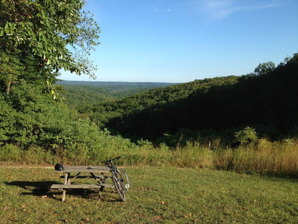 A scenic view of rolling hills and dense forests under a clear blue sky, with a picnic table and mountain bike in the foreground. A helmet rests on the table, suggesting a recent outdoor biking adventure. Brown County Park mountain bike trail.