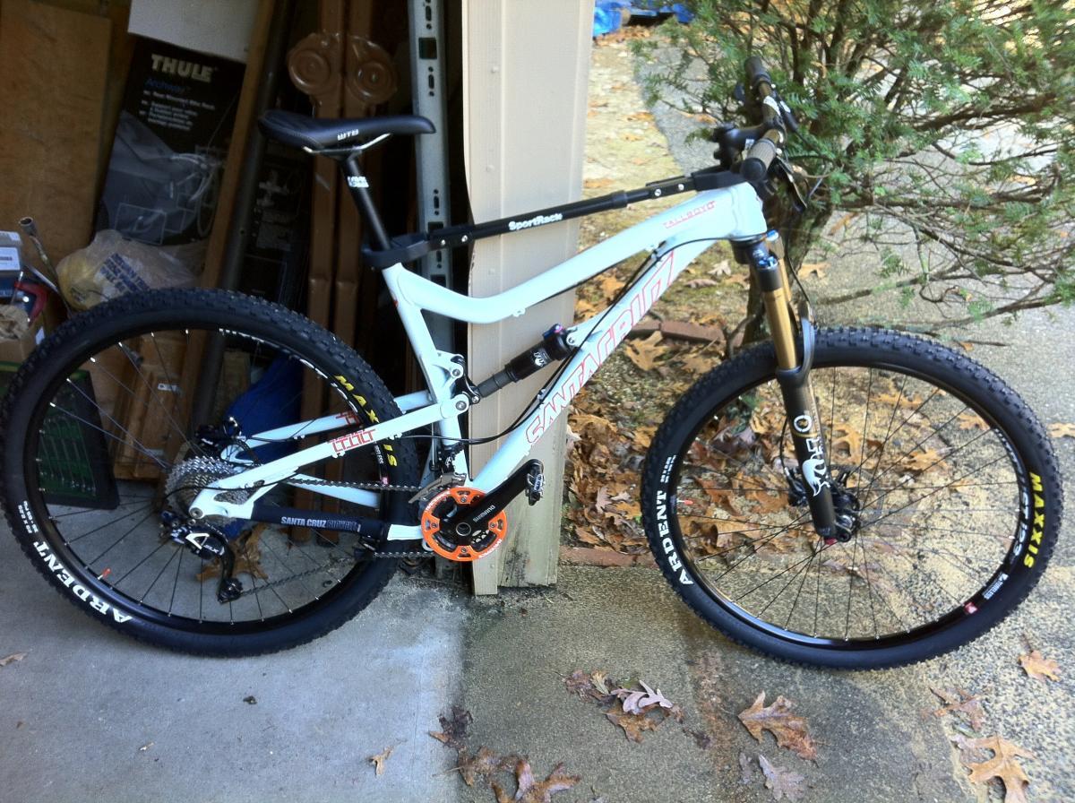 Santa Cruz Tallboy LT: A mountain bike with a white frame, featuring black and orange components, is positioned next to a garage wall. The bike has large tires and a sturdy build, ideal for off-road cycling. Surrounding the bike are fallen leaves and a glimpse of a bush, suggesting an outdoor setting.