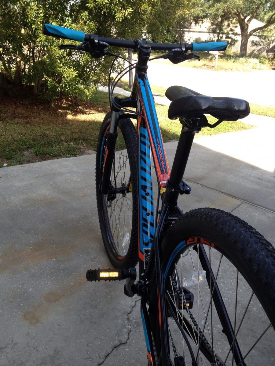 Specialized Hardrock Sport Disc 29er: Alt text: A close-up view of a mountain bike with a blue and orange design, showing the handlebars, seat, and part of the wheels. The bike is positioned on a concrete surface with greenery in the background.