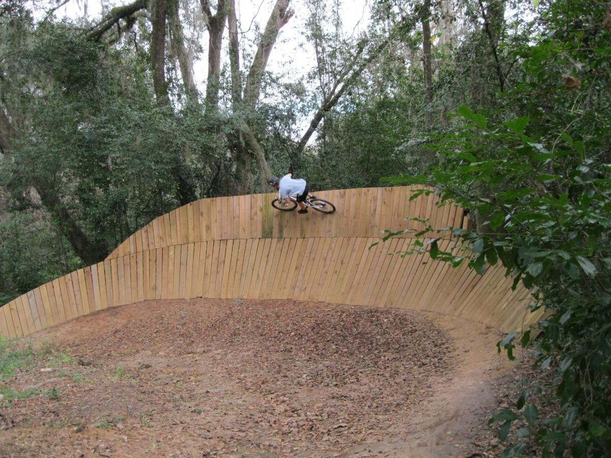 A person riding a bicycle on a wooden pump track, navigating a curved, elevated section surrounded by trees and foliage. The track features a smooth, banked wall designed for BMX or mountain biking, with a dirt surface covered in leaves and natural debris. Tom Brown / Lafayette Heritage Park mountain bike trail.
