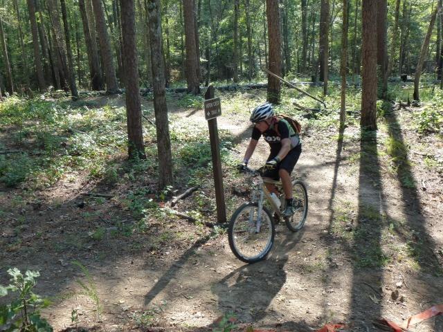 A person wearing a helmet and biking gear rides a mountain bike along a dirt trail in a wooded area, with tall trees and sunlight filtering through the leaves. A trail sign is visible nearby, indicating directions or information about the path. Lick Fork (Horn Creek) mountain bike trail.