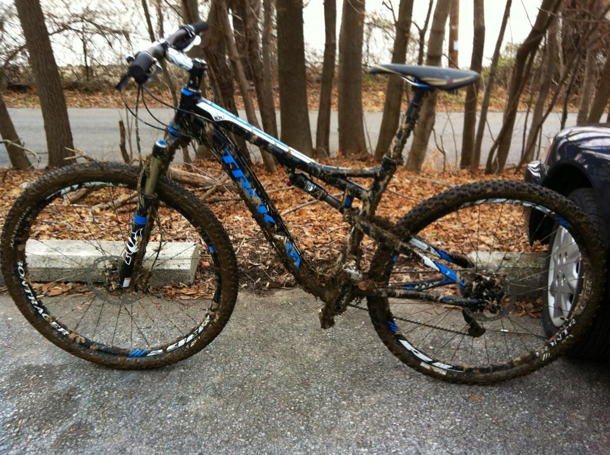 Trek Superfly 100 AL: A mountain bike covered in mud, parked on a gravel surface with trees in the background. The bike features a blue and black frame, thick tires, and is resting beside a gray vehicle. Brown leaves are visible on the ground, indicating an outdoor setting.