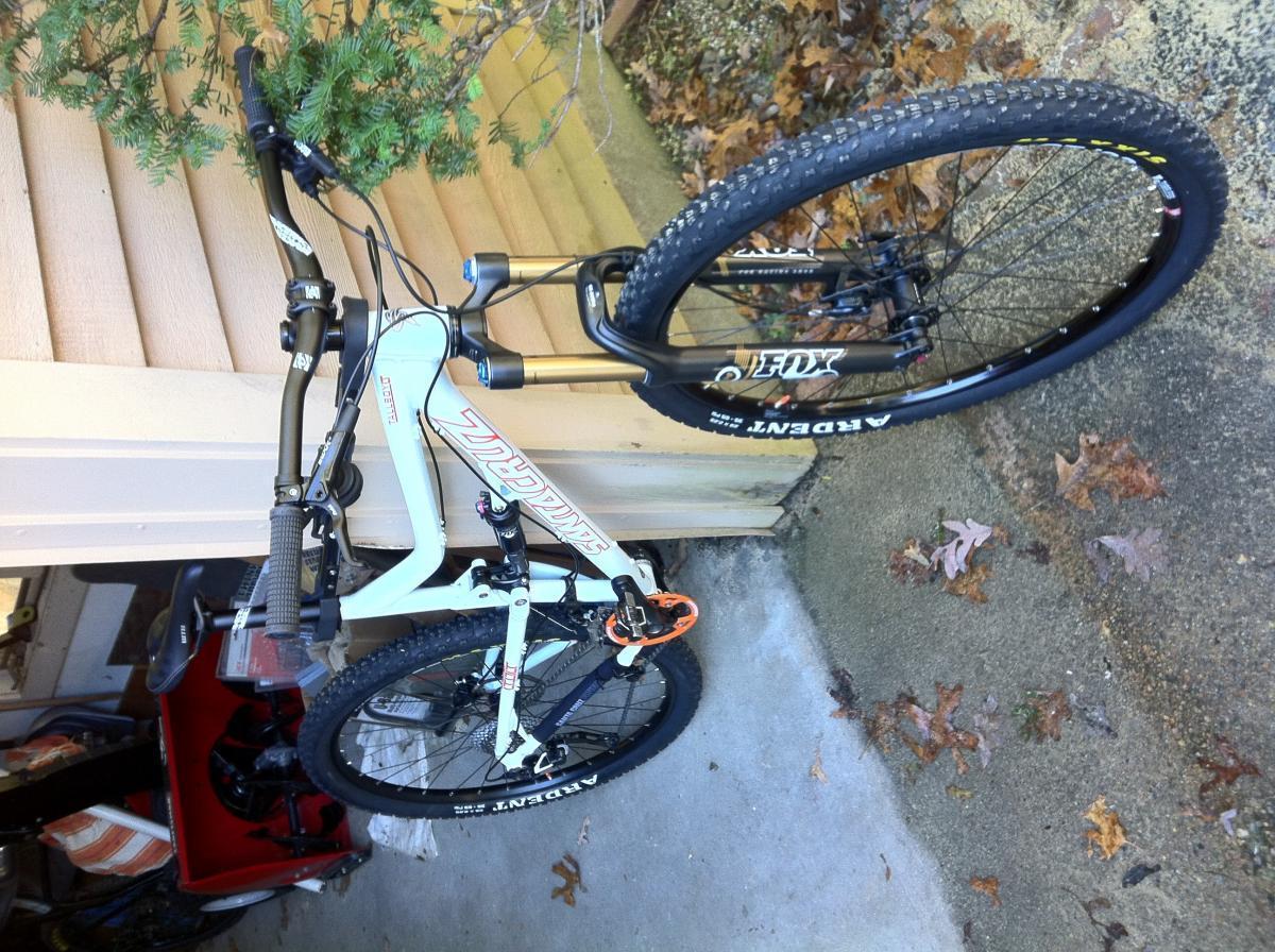 Santa Cruz Tallboy LT: A mountain bike leaning against a wooden surface, with the front wheel elevated. The bike features a white frame, black tires with prominent tread, and gold suspension components. In the background, there's a small red storage container and scattered leaves on the ground.