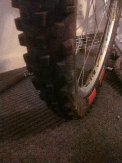 ITS 909: Close-up of a dirty mountain bike tire resting on a textured surface, showcasing the rugged tread pattern. The tire appears worn, indicating use on rough terrain.