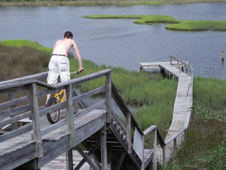 Monty Monty X-Lite: A person riding a yellow bike on a wooden walkway overlooking a marshy area with water and green grasses. The individual is shirtless and wearing white shorts, focusing on navigating the ramp. In the background, a serene landscape of water and marshland is visible.