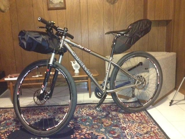 Motobecane Fly Team 29er Ti X0 Edition: A mountain bike with black saddle and handlebar bags, positioned in a cozy living room with wooden paneling and a patterned rug. A couch is visible in the background along with a small table and decor items.