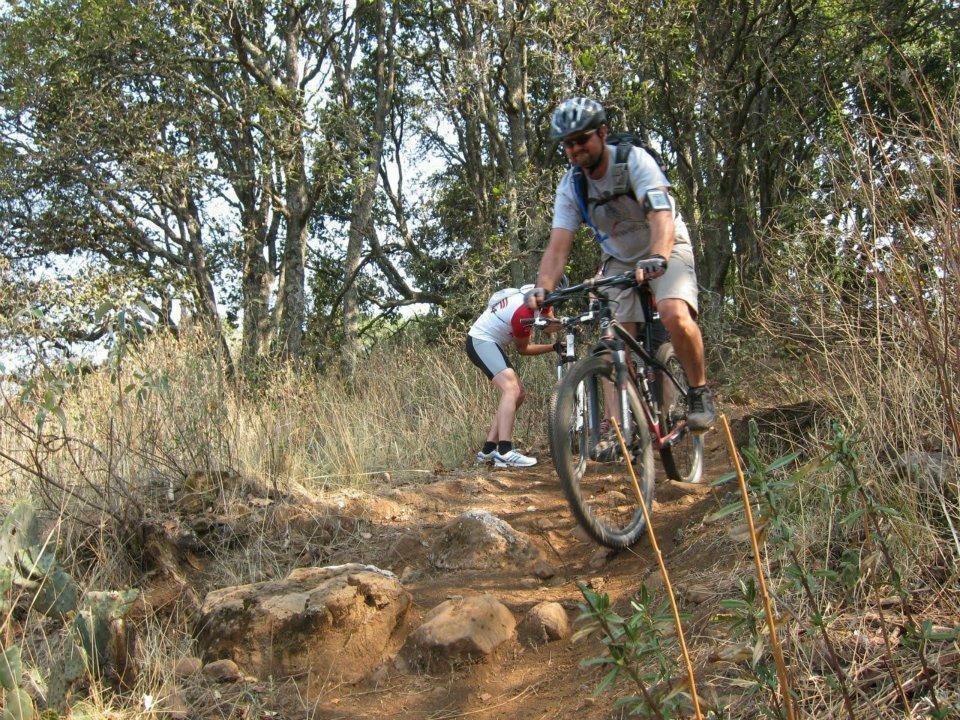 A man riding a mountain bike down a dirt trail in a wooded area, while another person kneels beside the path. The scene features greenery and rocky terrain typical of outdoor trails. Cerro Los Gallos mountain bike trail.
