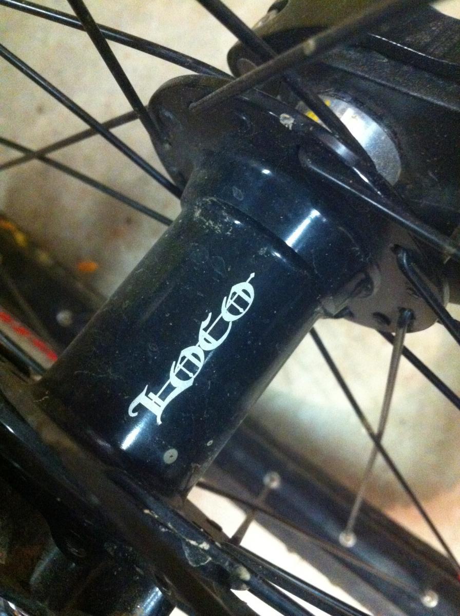 Forte Loco Front Wheel: Close-up of a bicycle hub with a "Loco" logo, surrounded by spokes and a slightly dusty surface. The background features part of the bike frame and floor.