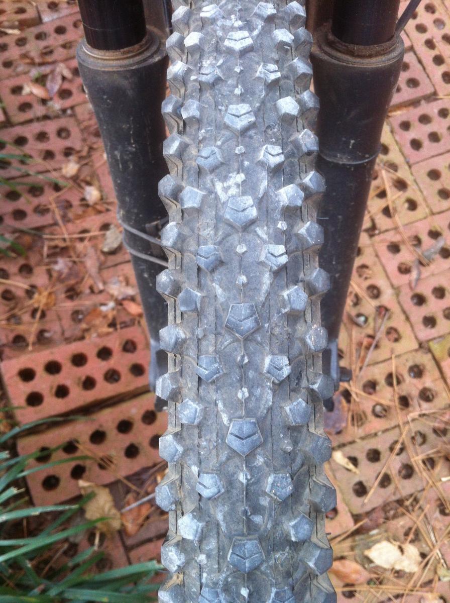 Maxxis Ignitor UST: Close-up view of a mountain bike tire with a rugged tread pattern, resting on a textured surface of bricks and grass. The tire shows signs of wear and dirt, indicating use in off-road conditions.