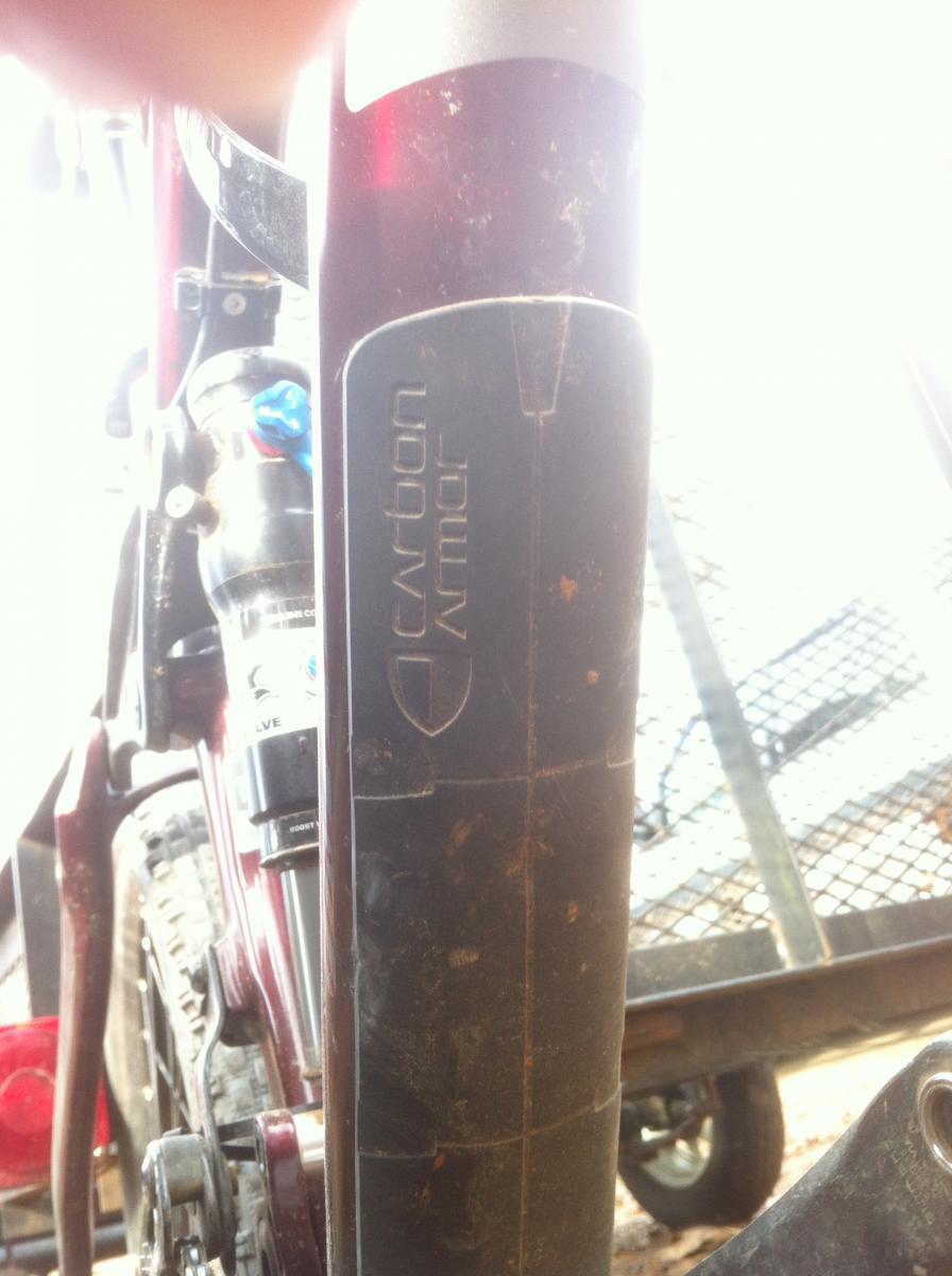 Trek Remedy 9.9: Close-up image of a bicycle fork featuring a "Carbon Armor" logo. The fork shows signs of dirt and wear, indicating usage. The background includes parts of a bike and a trailer.