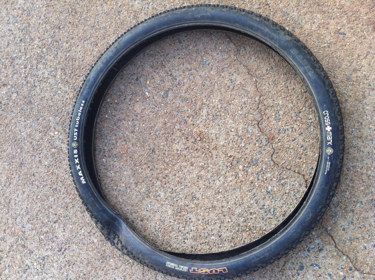 Maxxis Crossmark: A close-up view of a black bicycle tire lying on a textured concrete surface. The tire features markings that indicate it is a Maxxis CrossMark, designed for tubeless use.