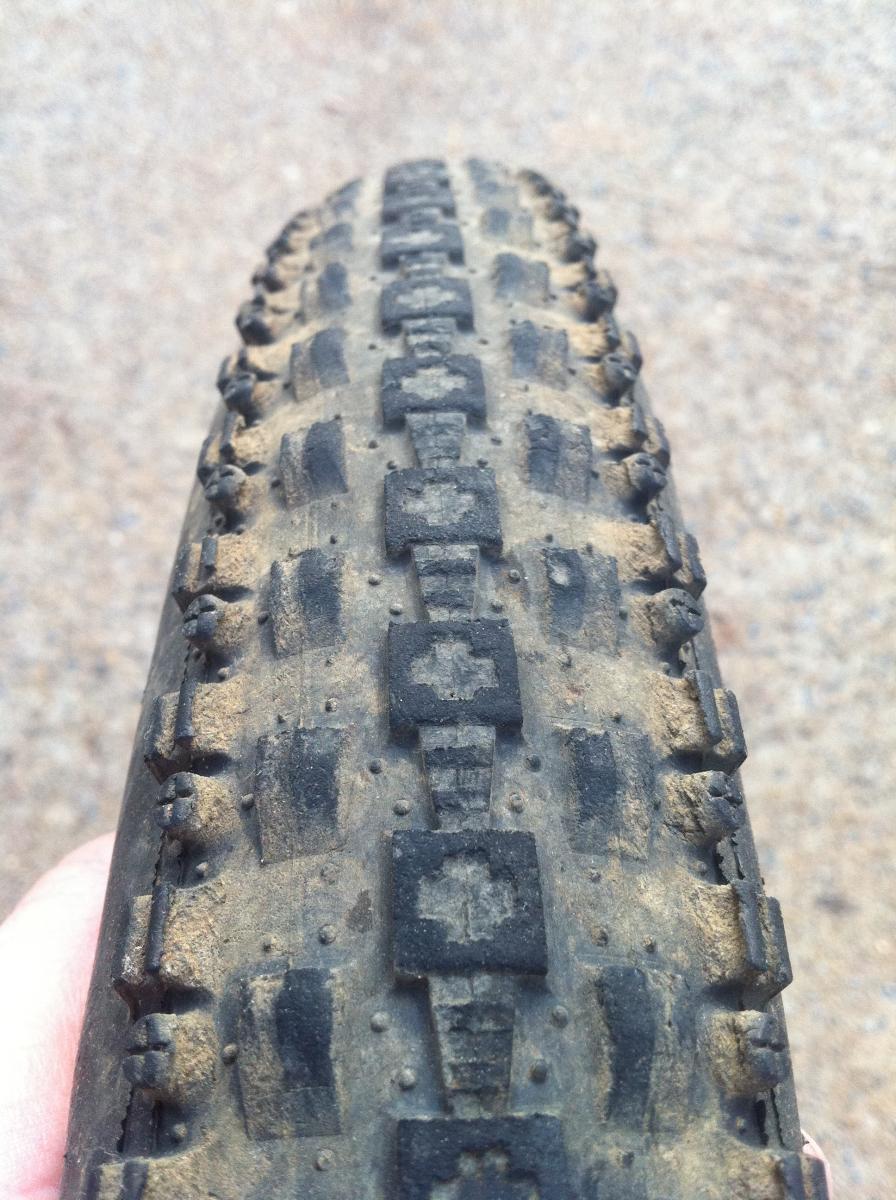 Maxxis Crossmark UST: Close-up view of a mountain bike tire showing its tread pattern, with visible dirt and wear, resting in a hand against a gravel background.