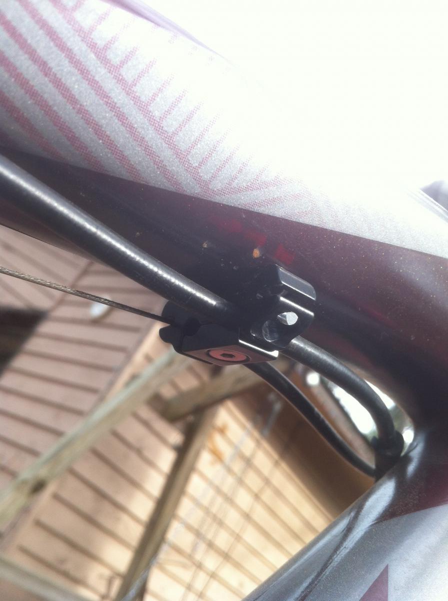 Trek Remedy 9.9: A close-up image of a bicycle's frame featuring a black cable guide attached to the top tube. The surrounding area shows a striped pattern on the bike frame, with a blurred wooden structure in the background.