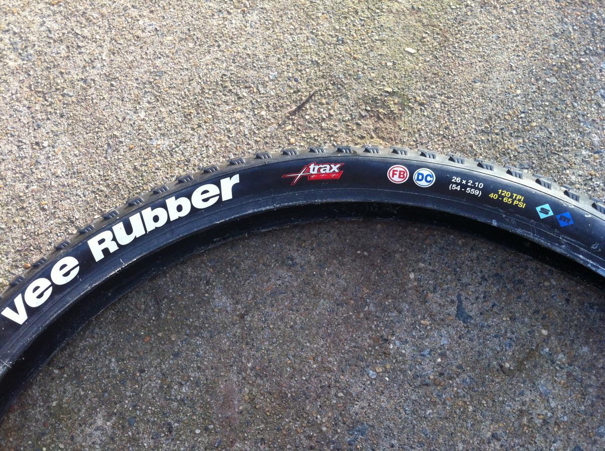Vee Rubber V8: A close-up view of a black bicycle tire with "Vee Rubber" printed in white letters. The tire has various markings indicating size (26 x 2.10), TPI (120), and PSI (40-65). The tire is resting on a textured surface.