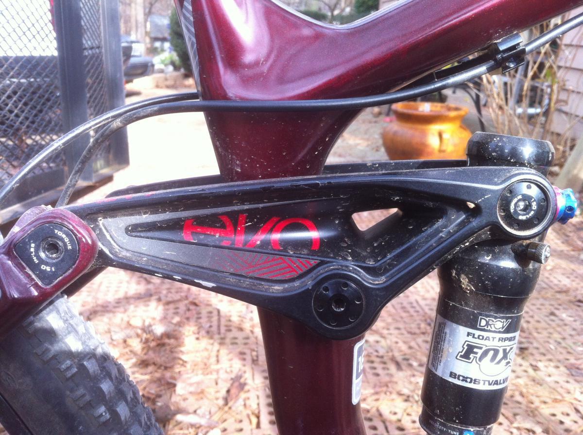 Trek Remedy 9.9: Close-up view of a mountain bike's rear suspension system, featuring a black and red linkage design. The image shows detailed components, including a shock absorber labeled "FOX Float RP3" and various adjustment points. The bike frame is maroon, with some dirt and mud visible, indicating off-road use. The background includes a blurred outdoor scene with exposed ground and a large pot.