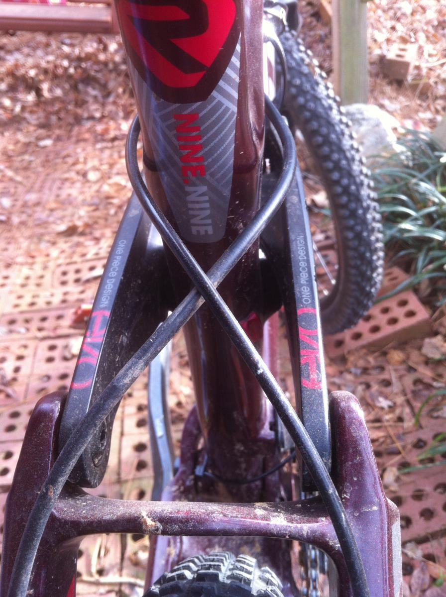 Trek Remedy 9.9: Close-up image of a bicycle frame, featuring a dark brown and red design with the text "NINE" on the top tube. The bike shows signs of use, with dust and dirt visible. A black cable is intertwined around the frame, and there are portions of the rear triangle that display the label "ONE". The background consists of earthy tones, including leaves and brick patterns.