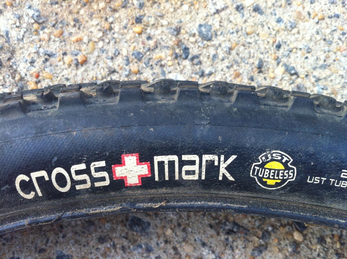 Maxxis Crossmark UST: Close-up of a bicycle tire with the text "cross mark" prominently displayed, along with a logo indicating "UST Tubeless" on its sidewall. The tire shows some wear and is lying on a rough gravel surface.