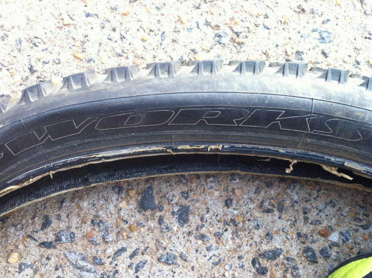 Specialized Fast Trak LK Pro: A close-up image of a damaged bicycle tire laying on a rough gravel surface, showing visible wear and tear along the edges and tread, with the brand name partially visible.