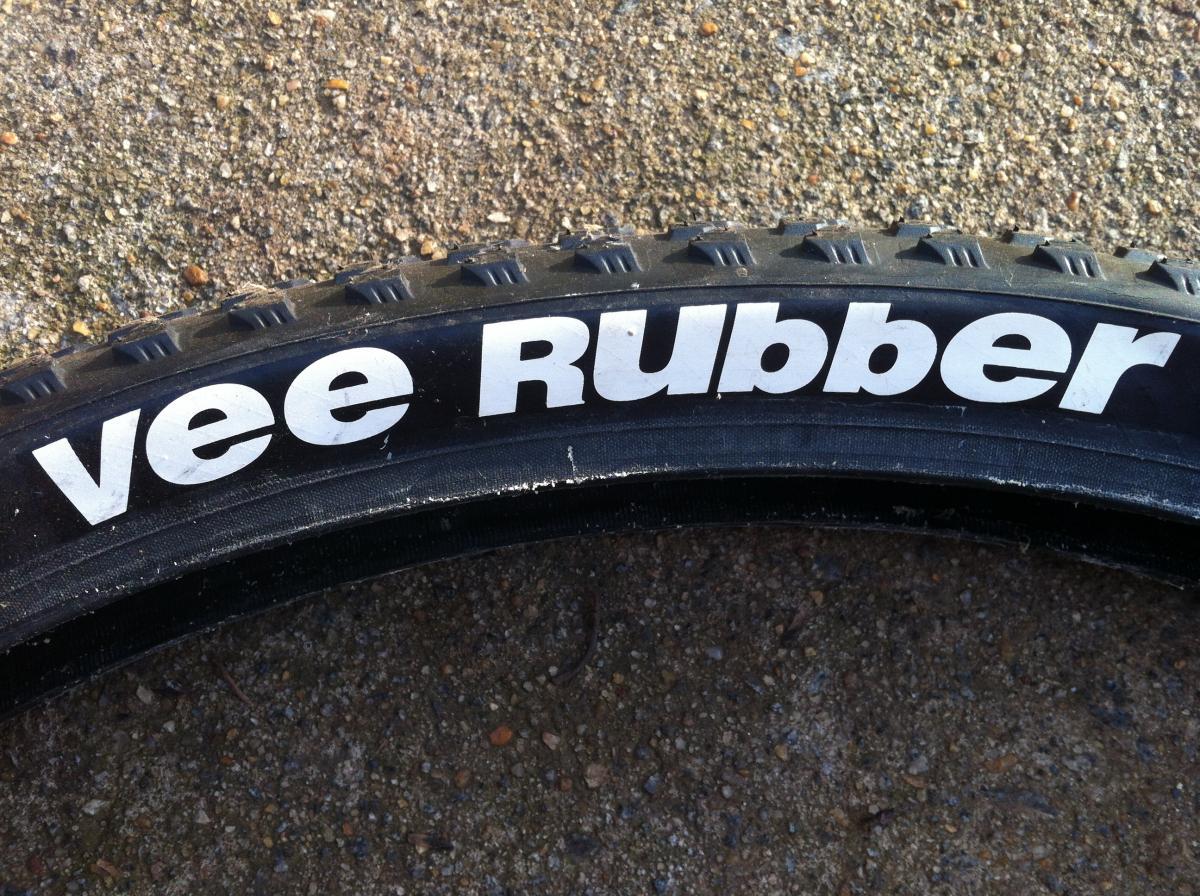 Vee Rubber V8: A close-up view of a bicycle tire with the text "vee Rubber" prominently displayed on the sidewall, set against a textured, gravel-like surface.