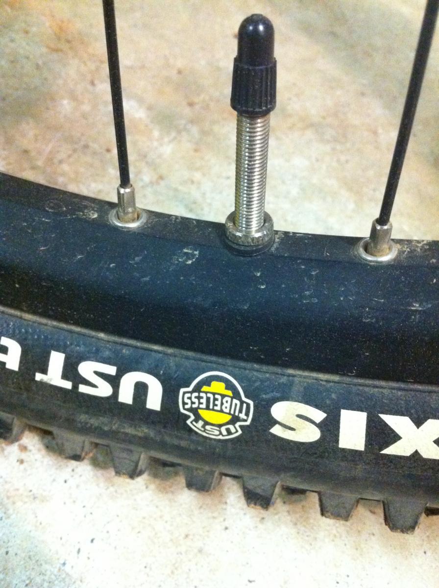 Forte Loco Front Wheel: Close-up view of a bicycle tire valve on a black tire with visible tread. The valve is a metallic Schrader type, partially threaded, alongside a logo for UST Tubeless. The surface of the tire shows signs of dirt, suggesting off-road use. The background is a concrete floor.
