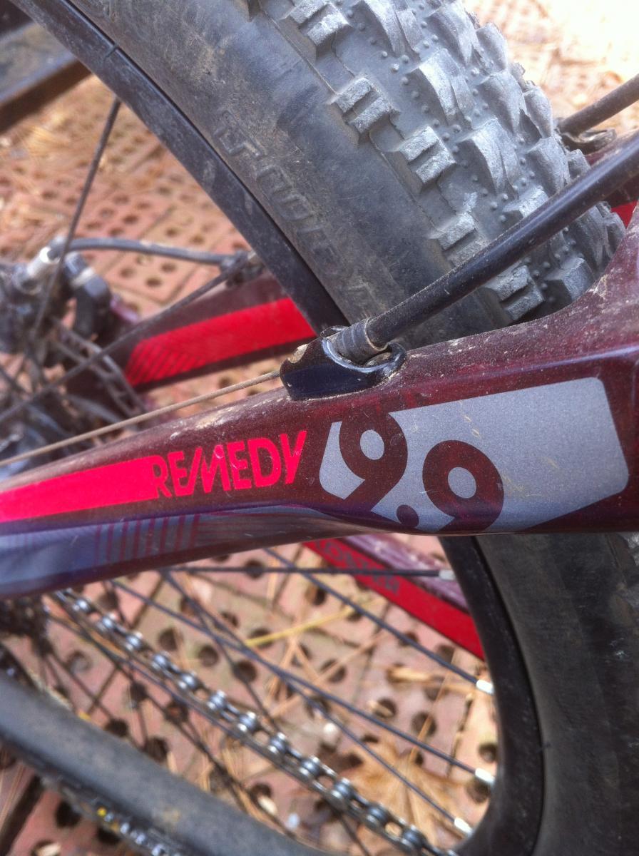 Maxxis Larsen TT: Close-up view of a mountain bike frame showing the "Remedy 9.0" branding, with a focus on the rear tire and chain. The bike is dusty, indicating recent outdoor use, and the background features a textured surface, likely a trail or path.