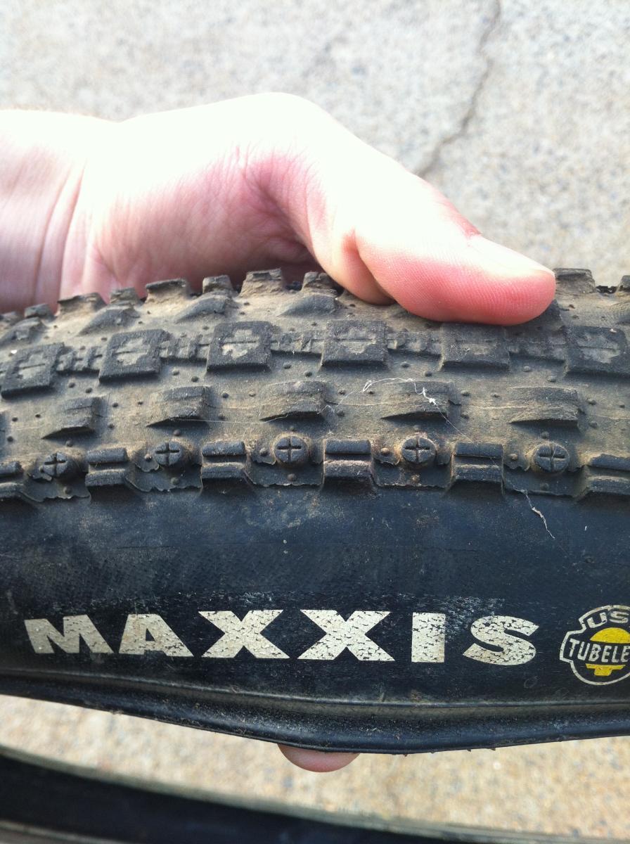 Maxxis Crossmark UST: A close-up view of a Maxxis bicycle tire being held by a person's hand, showing the tread pattern and brand logo. The tire has a dusty appearance, indicating use, and features raised sections and cross patterns on the tread.