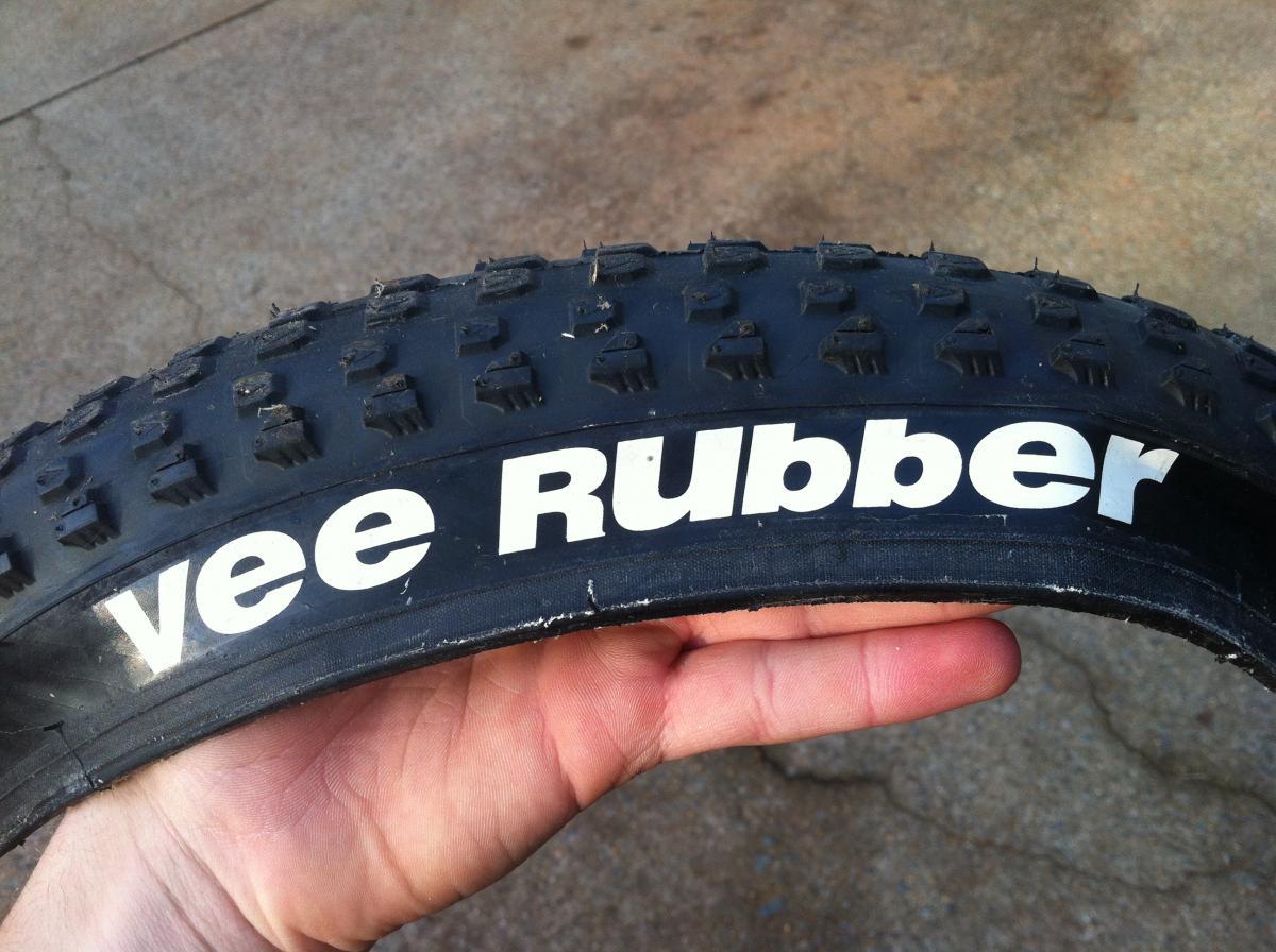 Vee Rubber V8: Close-up image of a black bike tire with a textured surface and the brand name "vee Rubber" prominently displayed in white lettering, held in a person's hand against a concrete background.