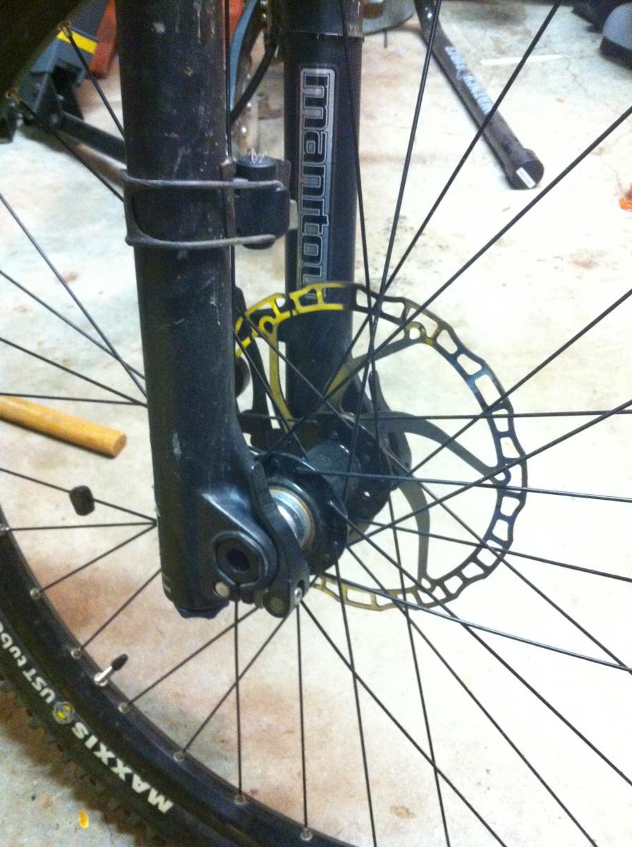 Forte Loco Front Wheel: Close-up view of a mountain bike front wheel, showing the fork, disc brake, and wheel hub. The bike has a black frame and components, with visible dirt on the fork, indicating use. The disc brake features a yellow accent, while the tire has "Maxxis" branding. The background is a garage setting with tools and equipment visible.