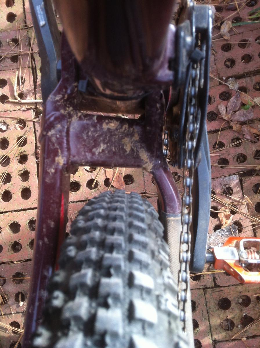 Trek Remedy 9.9: Close-up view of a mountain bike's rear wheel, showcasing the chain, gear mechanism, and partially visible frame. The tire features a rugged tread pattern, and there is dirt and mud visible on the bike components. The background consists of a textured surface with holes, likely indicating a trail or outdoor setting.