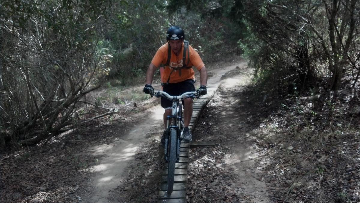 A person wearing a helmet and an orange shirt rides a mountain bike along a narrow wooden boardwalk, surrounded by greenery and trees. The path is partially covered in dirt and leaves, showing a natural outdoor setting for biking. Santos mountain bike trail.