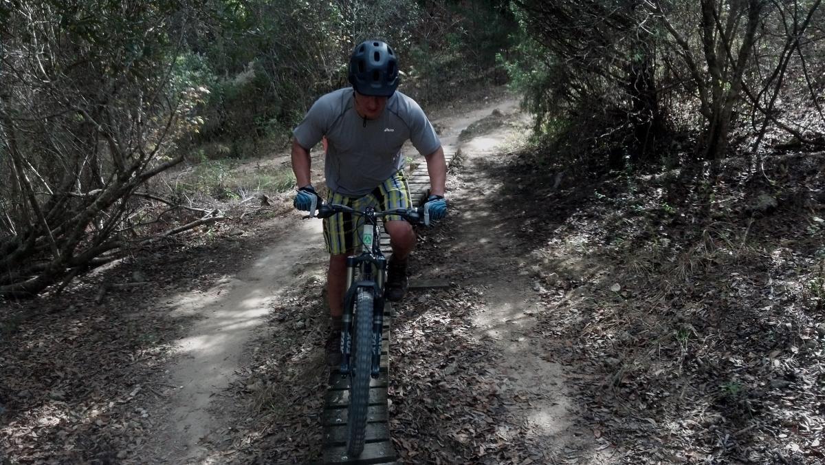 A person riding a mountain bike on a narrow, dirt trail surrounded by trees and foliage. The rider is wearing a helmet and protective gear, focused on navigating the path. The scene is set in a wooded area with a mix of sunlight and shade. Santos mountain bike trail.
