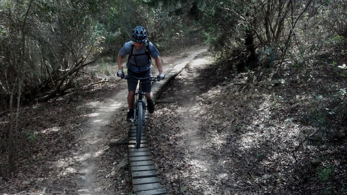 A mountain biker riding on a narrow wooden bridge through a forested trail, surrounded by greenery and fallen leaves. The cyclist is focused, wearing a helmet and gloves, while navigating the path. Santos mountain bike trail.