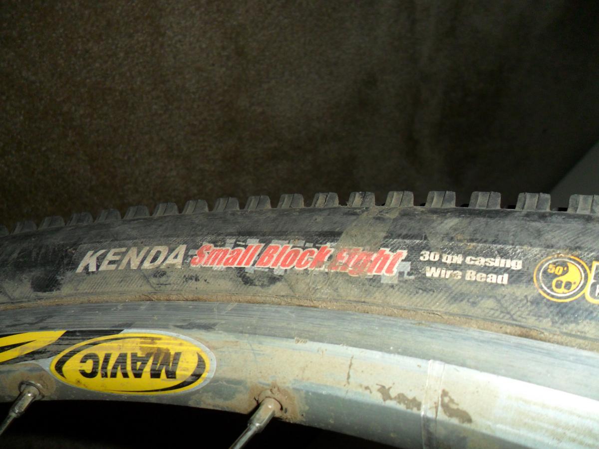 Kenda Small Block Eight: Close-up image of a Kenda Small Block Eight bicycle tire, featuring text that includes tire specifications such as "30 TPI casing" and "Wire Bead." The tire appears slightly dirty, indicating usage. The image also shows part of the rim with a NAYAK logo.