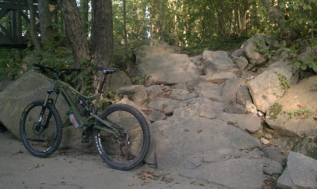 Santa Cruz Nickel: A mountain bike parked next to a rugged rocky slope surrounded by trees and foliage, with a pathway leading through the natural environment.