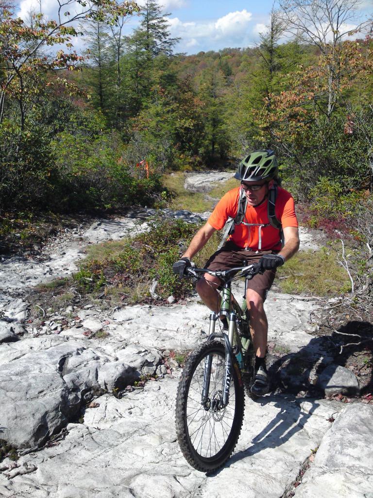 Santa Cruz Nickel: A cyclist in an orange shirt and helmet navigates a rocky trail surrounded by trees and greenery. The rider appears focused as he balances on his mountain bike, set against a backdrop of blue sky and distant hills.