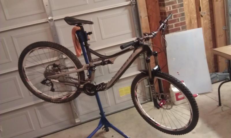 Specialized Stumpjumper FSR Comp 29: A mountain bike mounted on a repair stand in a garage, with a garage door and wooden walls in the background. The bike is upside down, showing its wheels and frame, and has an orange cloth hanging from the seat post. A table with various items can be seen in the background.