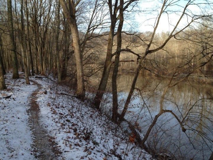 A snowy pathway winds alongside a tranquil river, bordered by bare trees and winter foliage. The scene captures a serene winter landscape with gentle sunlight illuminating the atmosphere. Versailles State Park mountain bike trail.