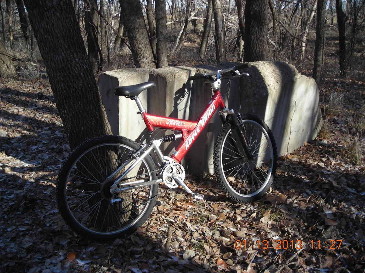 A red mountain bike leaning against a tree and a concrete block in a wooded area, with fallen leaves scattered on the ground. The scene is illuminated by natural light, indicating a cool day. ESU Trail mountain bike trail.