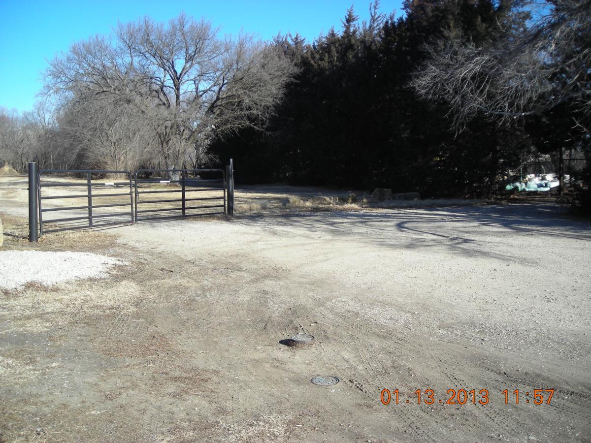 A gravel road leading to a black metal gate, surrounded by bare trees on either side. The ground is dry, and the scene is under a clear blue sky. The image is timestamped January 13, 2013, at 11:57 AM. ESU Trail mountain bike trail.