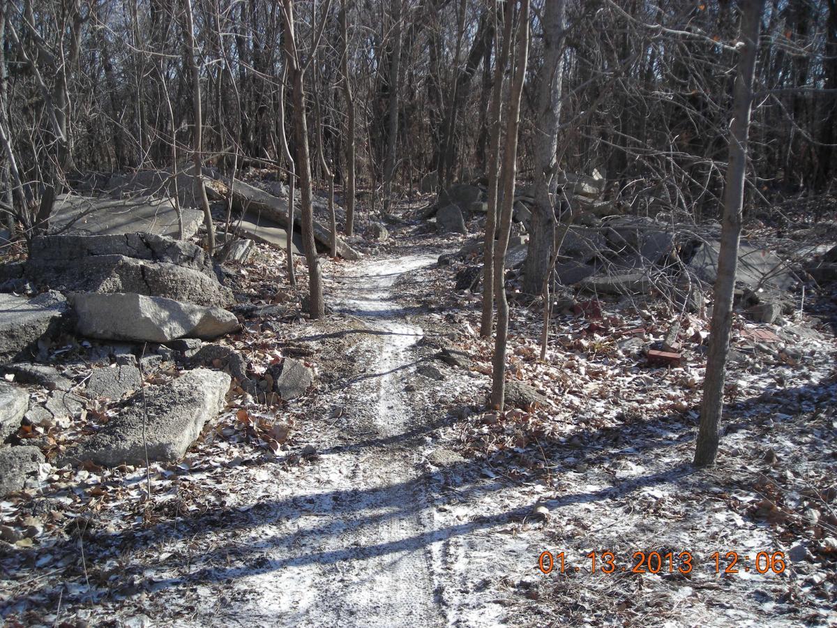 A narrow, winding dirt trail meanders through a forested area, flanked by bare trees and scattered rocks. The ground is covered with fallen leaves and a light dusting of snow, suggesting a chilly winter day. Sunlight filters through the branches, casting a mix of shadows on the path. ESU Trail mountain bike trail.
