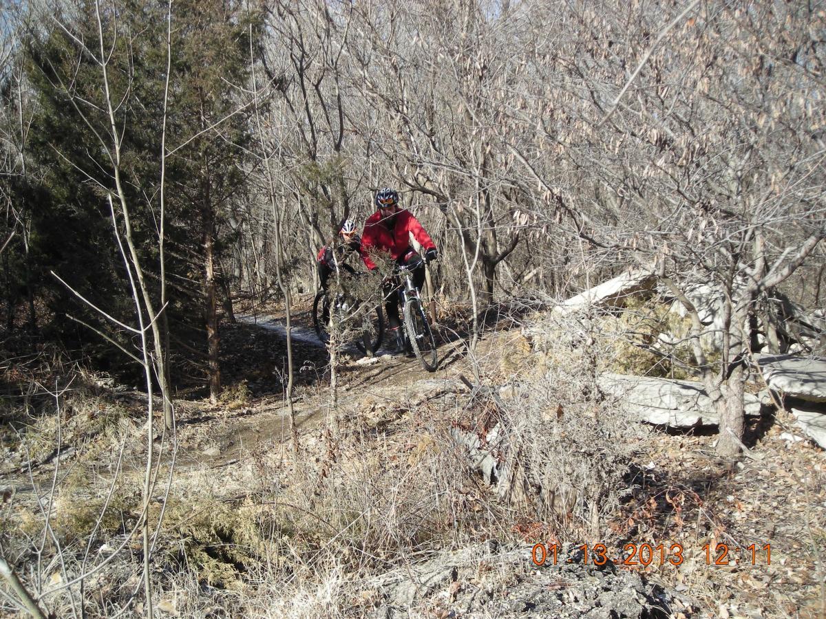 Two mountain bikers navigated a narrow, winding trail through a sparse, leafless forest. The riders are dressed in winter biking gear, with one in a red jacket. The terrain includes patches of dirt and rocks, surrounded by bare trees and underbrush. ESU Trail mountain bike trail.