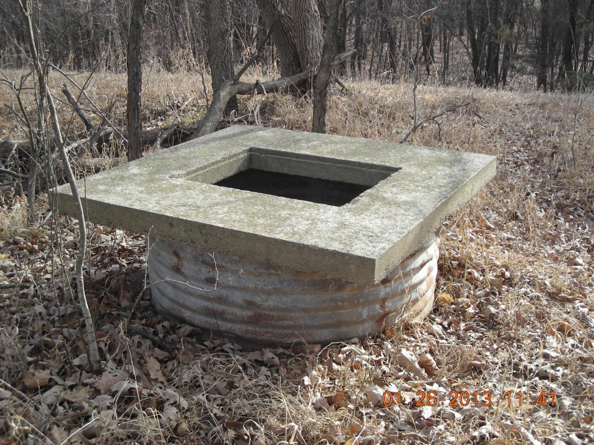 A concrete well cover resting on a rusted metal pipe, surrounded by dry grass and trees in a natural setting. The surface of the well cover is slightly weathered, and the date "01/26/2013 11:41" is visible in the corner. ESU Trail mountain bike trail.