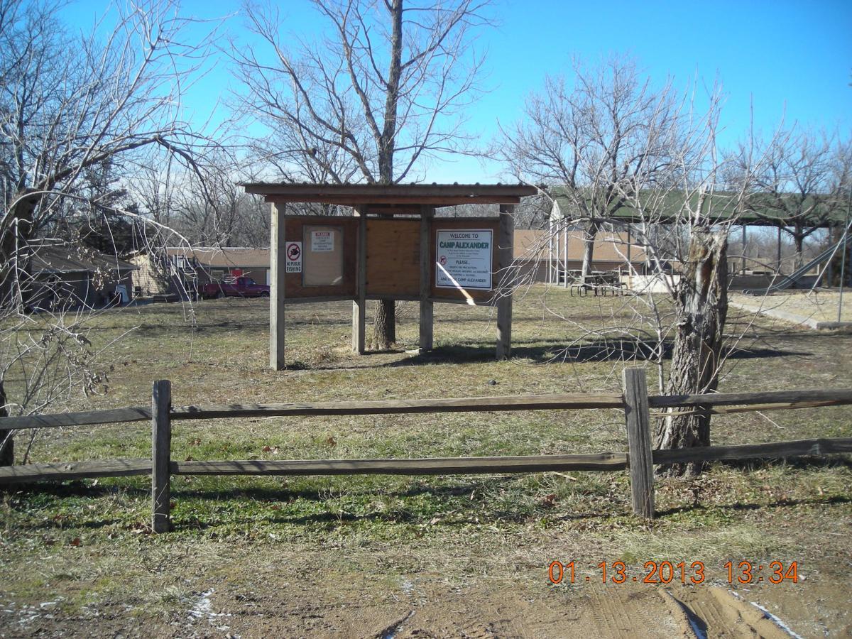 Image of a sign at Camp Alexander, surrounded by a grassy area and bare trees. The sign displays "Welcome to Camp Alexander" and additional informational notices. In the background, there are buildings and a swing set visible, with a wooden fence in the foreground. The sky is clear and blue, indicating a sunny day. Camp Alexander Trails mountain bike trail.