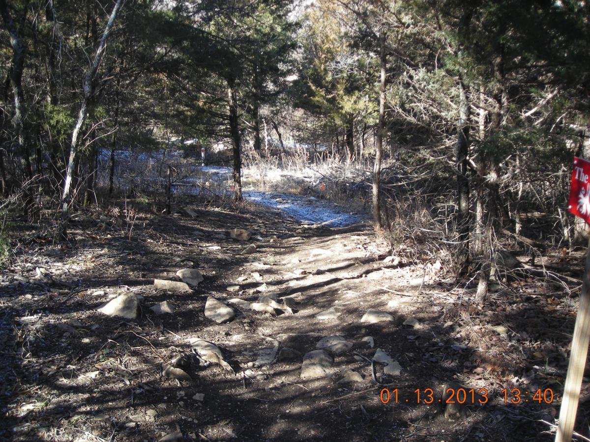 A rocky trail winding through a forested area, flanked by trees and underbrush. The path is natural with scattered stones, leading towards a wooded section with a hint of snow on the ground. A red signpost indicating a trail marker is visible on the right. Camp Alexander Trails mountain bike trail.