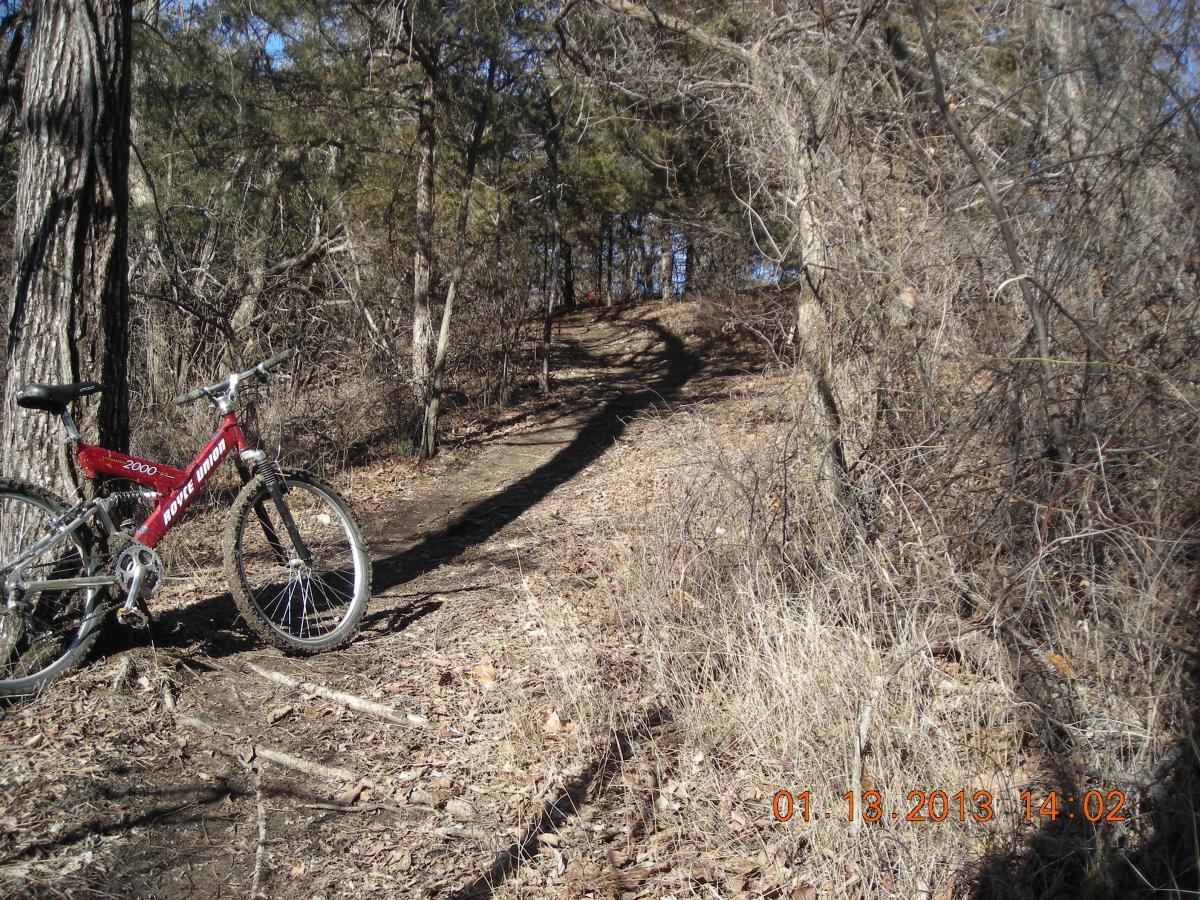 A red mountain bike rests on a dirt path surrounded by trees and bushes in a wooded area. The trail curves upwards in the background, indicating a hilly terrain. Sunlight filters through the branches, creating dappled shadows on the ground. The image captures a serene moment in nature, inviting outdoor exploration. Camp Alexander Trails mountain bike trail.